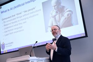 Man in suit and bowtie talking to audience, unseen, with presentation of AI behind him