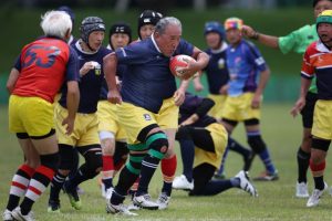 Older rugby players in protective headgear run after the ball during a match on a grass field