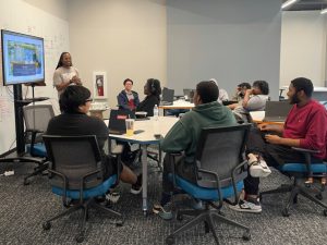 A class of young adults sit at their desks while an adult stands at the side of the class in front of a TV screen.