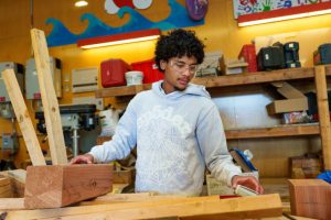 A person with curly hair and safety glasses is working with wooden beams in a workshop filled with tools and storage shelves.