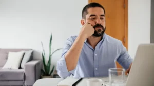 A short-bearded man in a business shirt sitting at a desk in front of a laptop, leaning on his elbow and looking bored.
